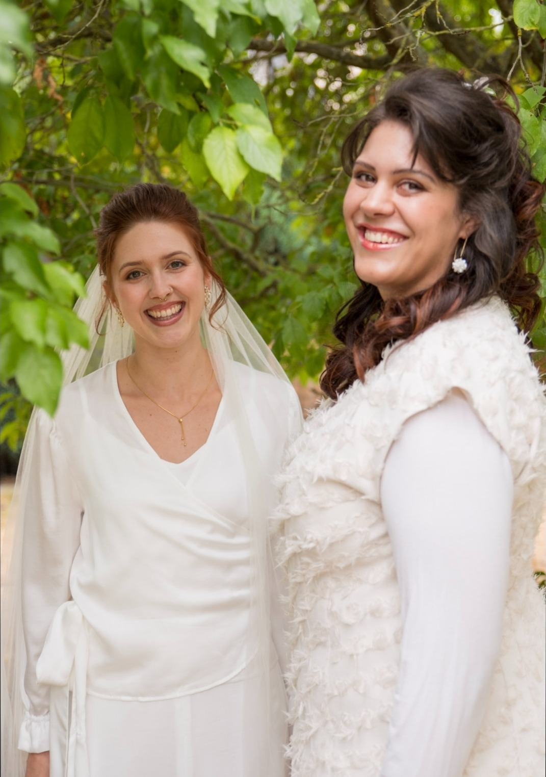 Zwei fertig gestylte Bräute, eine mit Schleier, die andere mit Locken und floralen Ohrringen, lachen nebeneinander im Grünen