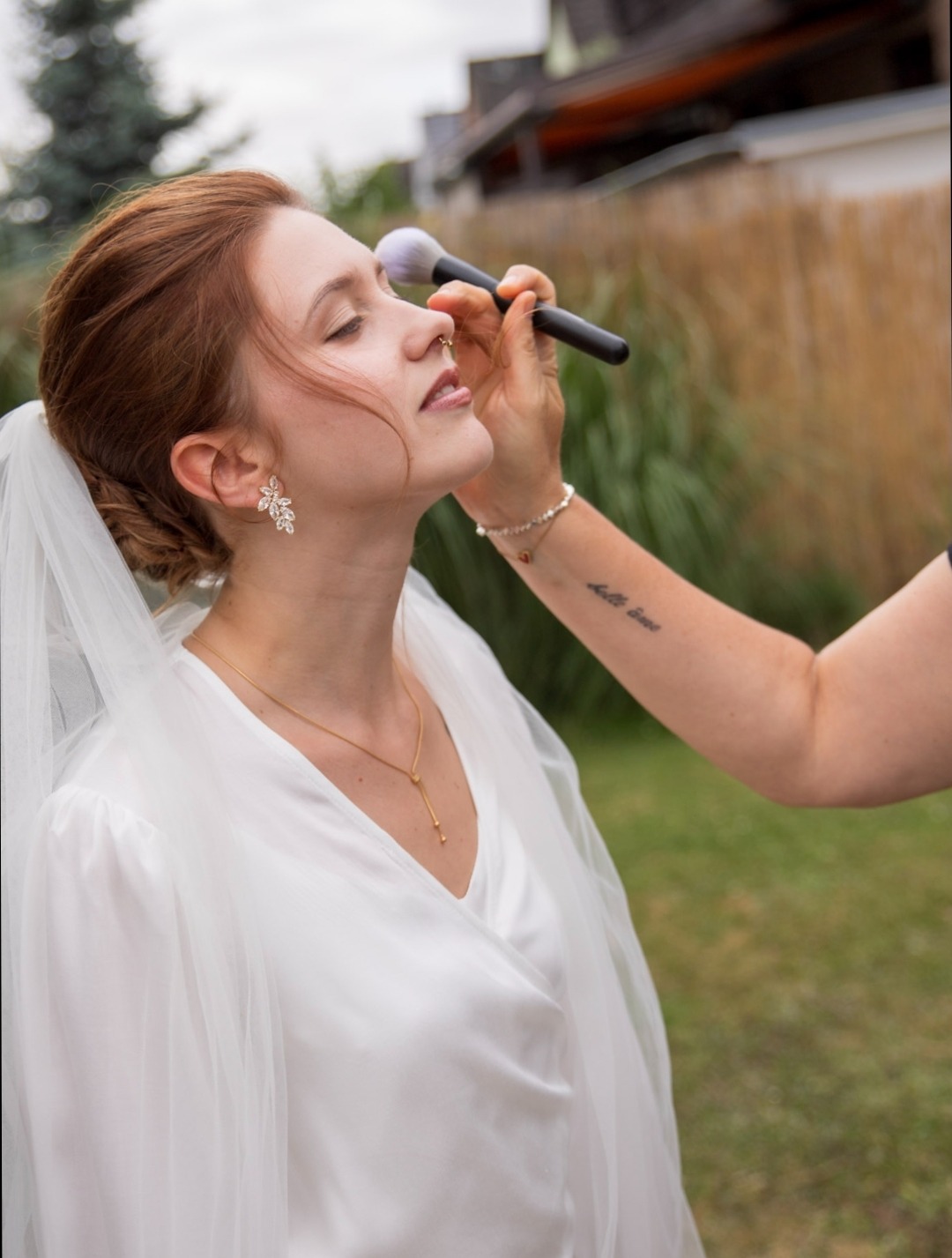Profil einer Braut mit Chignon und Schleier, während eine Stylistenhand mit einem großen Puderpinsel den letzten Touch aufträgt